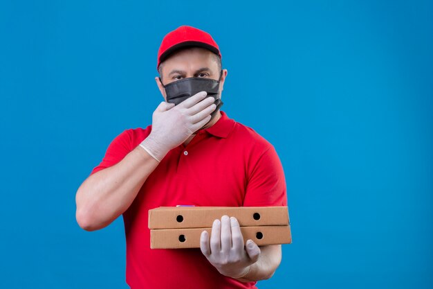 Delivery man wearing red uniform and cap in facial protective mask holding pizza boxes looking surprised covering mouth with hand over blue wall
