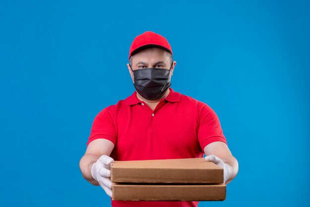 Delivery man wearing red uniform and cap in facial protective mask holding pizza boxes stretching out to camera looking confident over blue wall