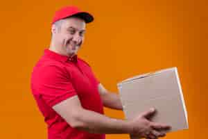 Free photo delivery man wearing red uniform and cap giving cardboard box to a customer with confident smile over isolated orange wall