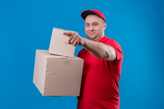 Delivery man wearing red uniform and cap holding cardboard boxes looking aside and pointing to something with index finger standing over isolated blue space