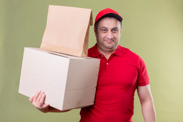 Delivery man wearing red uniform and cap holding cardboard boxes looking confident smiling over isolated green wall