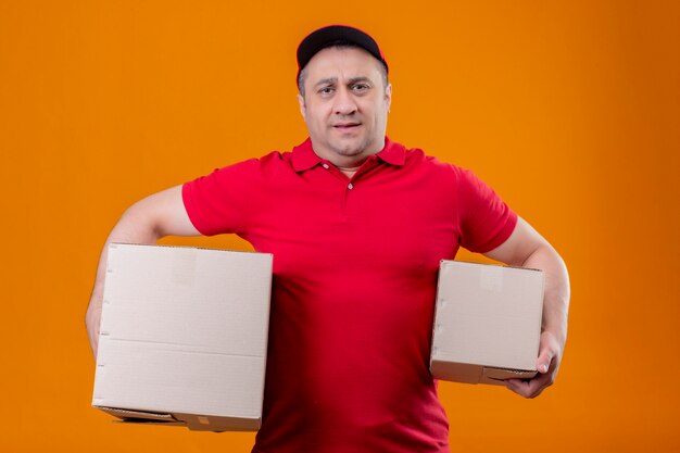 Работник службы доставки, одетый в красную форму и кепку с картонными коробками, выглядящий перегруженным и уставшим над оранжевой стеной