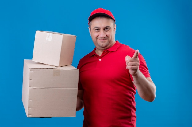Доставщик в красной форме и кепке держит картонные коробки, указывая на что-то указательным пальцем, уверенно и счастливо улыбаясь, стоя над синим пространством