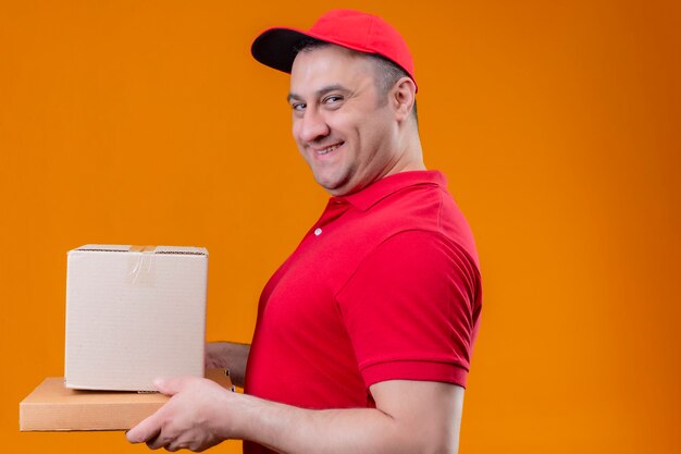 Доставщик в красной форме и кепке держит картон, стоя сбоку, уверенно улыбаясь