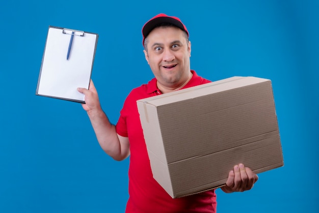 Доставщик в красной форме и кепке держит большую картонную коробку с буфером обмена и выглядит удивленным, стоя над изолированным синим пространством