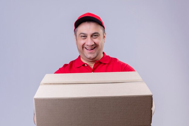 Fattorino che indossa l'uniforme rossa e cappuccio che giudicano sorridere grande della scatola di cartone amichevole con il fronte felice sopra la parete bianca isolata