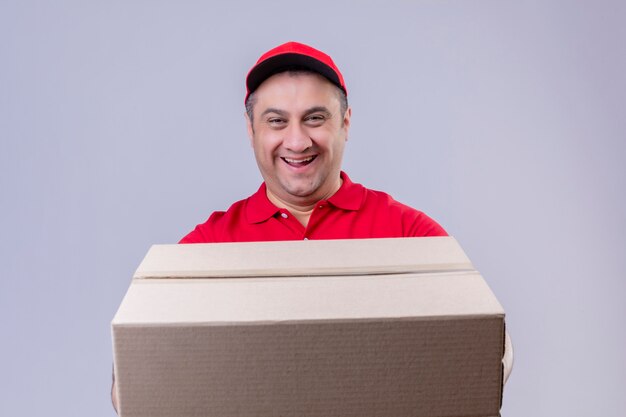 Delivery man wearing red uniform and cap holding large cardboard box smiling friendly with happy face over isolated white wall