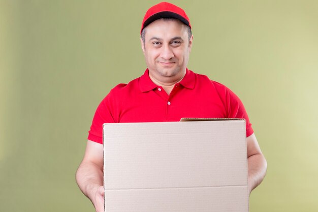 Доставщик в красной форме и кепке держит большую картонную коробку, дружелюбно улыбаясь со счастливым лицом, стоящим над изолированным зеленым пространством