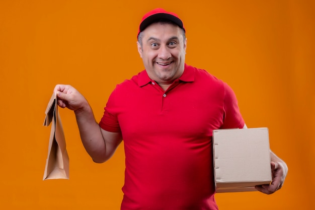 Delivery man wearing red uniform and cap holding paper package and cardboard box looking surprised and happy over orange wall