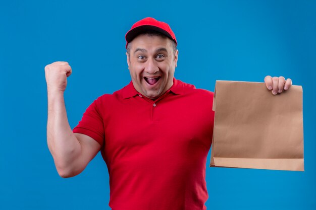 Delivery man wearing red uniform and cap holding paper package exited and happy raising fist after a victory over isolated blue wall