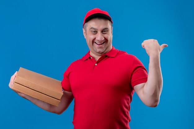 빨간 유니폼을 입고 배달 남자 격리 된 파란색 벽에 승리 후 주먹을 올리는 행복 한 얼굴로 웃고 피자 상자를 들고 모자