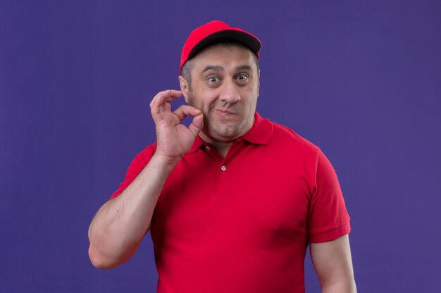 Delivery man wearing red uniform and cap making silence gesture with hand doing like closing mouth with a zipper sanding over purple space