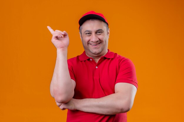 Доставка человек в красной форме и кепке положительно и счастливо указывая указательным пальцем в сторону, весело улыбаясь над оранжевой стеной
