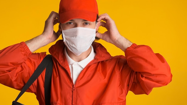 黄色の背景に分離された自信を持って見える赤い制服を着たフェイスマスクを持った配達人男性の宅配便は働く準備ができています安全第一
