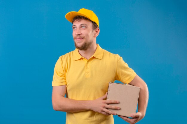 delivery man in yellow polo shirt and cap holding box package in hands smiling friendly looking up standing on blue