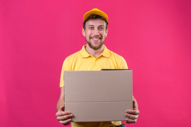 delivery man in yellow polo shirt and cap holding box package looking confident smiling cheerfully standing on isolated pink