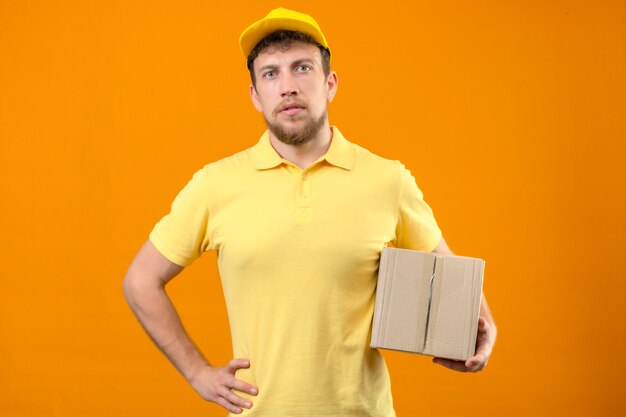 노란색 폴로 셔츠와 모자 상자 패키지를 들고 배달 남자 격리 된 오렌지에 서 혼란 찾고