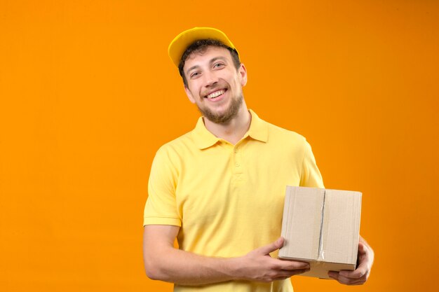 курьер в желтой рубашке поло и кепке держит коробку с пакетом, весело улыбаясь, глядя в камеру, стоящую на оранжевом