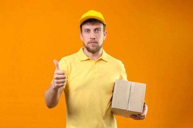 delivery man in yellow polo shirt and cap holding cardboard box looking confident showing thumb up standing on orange