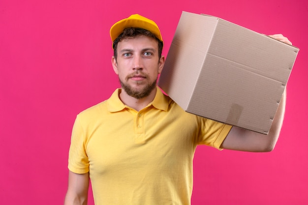 delivery man in yellow polo shirt and cap holding cardboard box on shoulder looking at camera with serious face standing on isolated pink