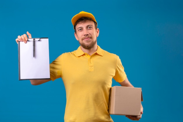 delivery man in yellow polo shirt and cap holding cardboard box showing clipboard with blanks smiling friendly on blue