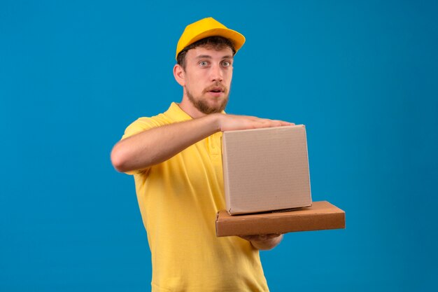 delivery man in yellow polo shirt and cap holding cardboard boxes in hands looking at camera surprised standing on blue