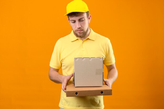 delivery man in yellow polo shirt and cap holding cardboard boxes looking sad with unhappy face standing on isolated orange