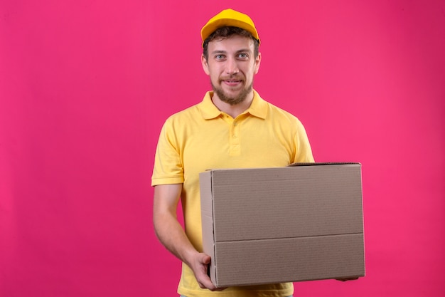 delivery man in yellow polo shirt and cap holding large cardboard box looking confident smiling friendly with happy face standing on isolated pink