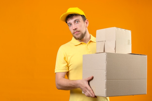 delivery man in yellow polo shirt and cap holding large cardboard boxes looking at camera with serious face standing on orange