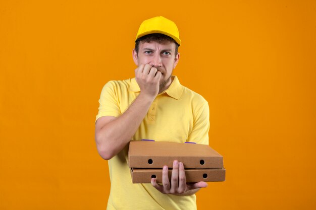 delivery man in yellow polo shirt and cap holding pizza boxes looking worried nervous biting fingernails standing on isolated orange