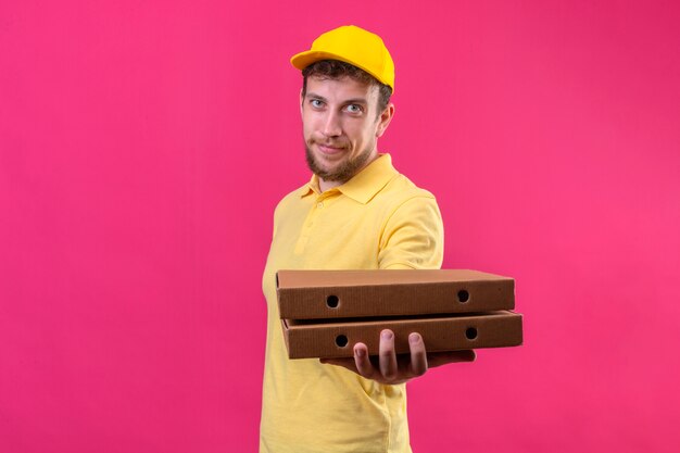 delivery man in yellow polo shirt and cap holding pizza boxes stretching out to customer with friendly smile on face on isolated pink