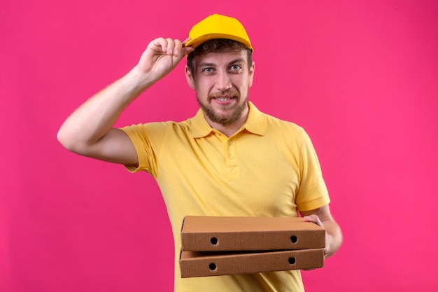 доставщик в желтой рубашке поло и кепке держит коробки для пиццы, трогая его кепку, глядя в камеру с дружелюбной улыбкой, стоя на розовом