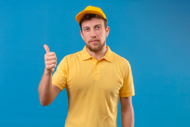 Курьер в желтой рубашке поло и кепке выглядит уверенно, показывает палец вверх, стоя на изолированном синем