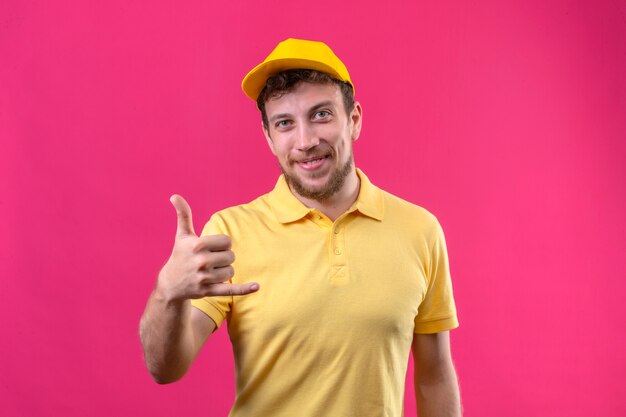 delivery man in yellow polo shirt and cap making call me gesture looking confident smiling friendly on pink