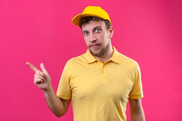노란색 폴로 셔츠와 격리 된 분홍색에 자신감이 미소로 측면에 검지 손가락으로 가리키는 모자 배달 남자