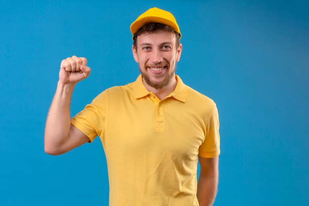 delivery man in yellow polo shirt and cap raising fist with happy face winner concept standing on blue