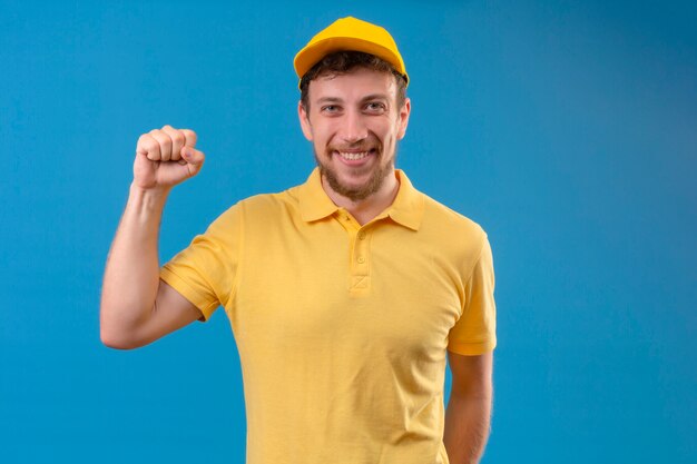 доставщик в желтой рубашке поло и кепке, поднимающей кулак с концепцией победителя счастливого лица, стоящей на синем