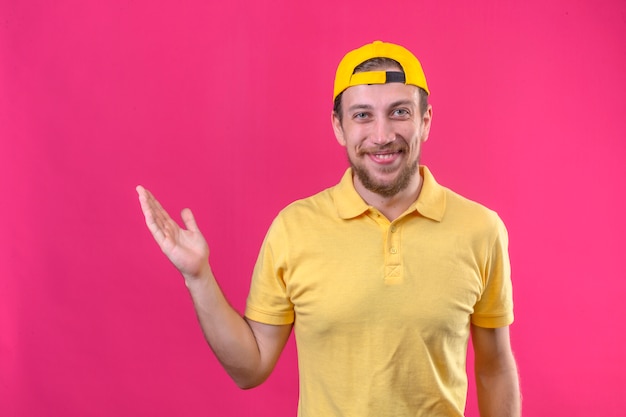 delivery man in yellow polo shirt and cap smiling cheerfully presenting and pointing with palm of hand standing on isolated pink