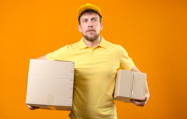 Delivery man in yellow polo shirt and cap standing with box package and large cardboard box with serious face without smile on orange