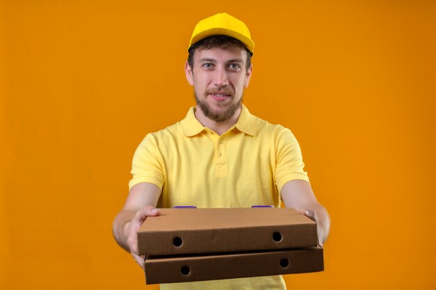 курьер в желтой рубашке поло и кепке протягивает коробки из-под пиццы, дарит покупателю дружелюбно улыбающийся на изолированном апельсине