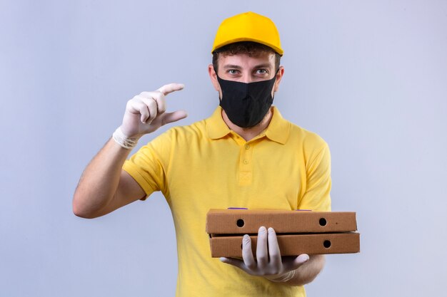 курьер в желтой рубашке поло и кепке, одетый в черную защитную маску, держит коробки для пиццы, жестикулируя руками, показывая символ меры знака небольшого размера на изолированном белом