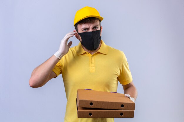 Доставщик в желтой рубашке поло и кепке, одетый в черную защитную маску, держит слабослышащие коробки из-под пиццы, стоя с пальцем возле уха на изолированном белом
