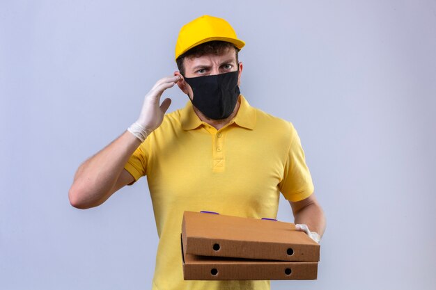 delivery man in yellow polo shirt and cap wearing black protective mask holding pizza boxes hard of hearing standing with finger near ear on isolated white