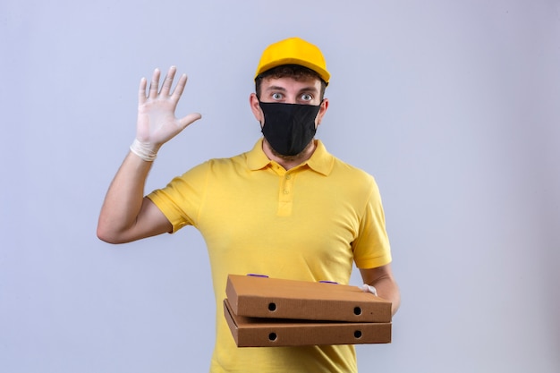 Fattorino in maglietta polo gialla e berretto che indossa una maschera protettiva nera con scatole per pizza che mostrano il numero cinque con la mano che sembra sorpreso in piedi su bianco