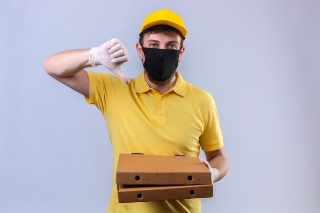 delivery man in yellow polo shirt and cap wearing black protective mask holding pizza boxes showing thumbs down standing on isolated white