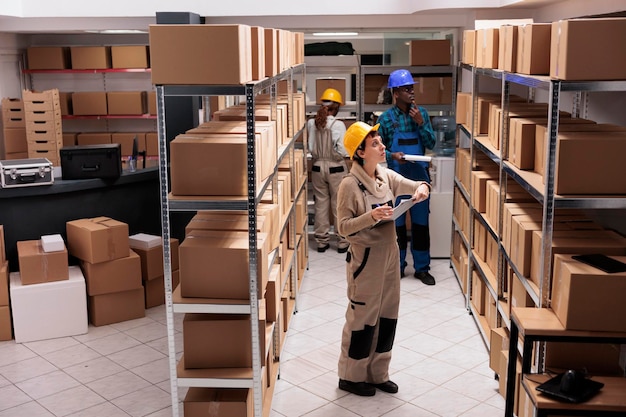 配達サービス員が倉庫で顧客の小包を検索し、クリップボードを保持しています。工場の倉庫で商品を監督する、制服のオーバーオールを着た多様な倉庫従業員