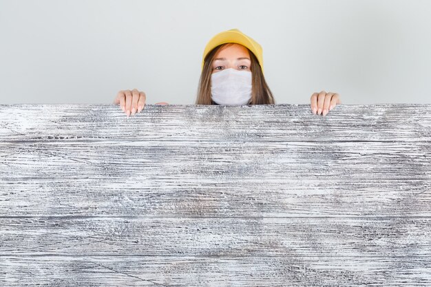 Delivery woman in cap, mask peeking behind wooden board