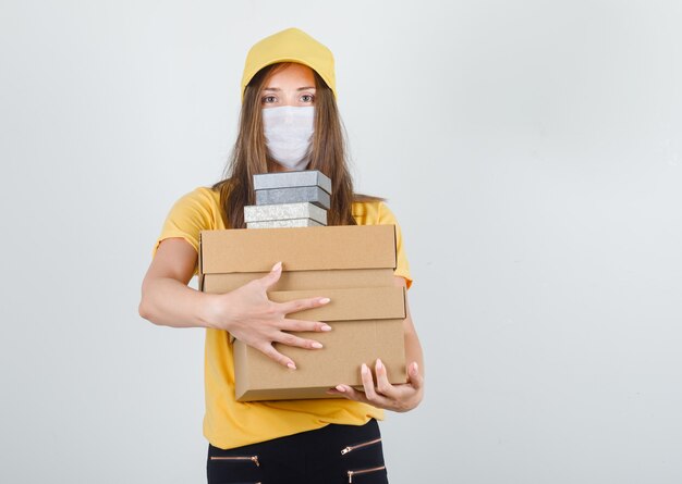 Женщина-доставщик обнимает коробки в футболке, штанах и кепке, маске и выглядит весело