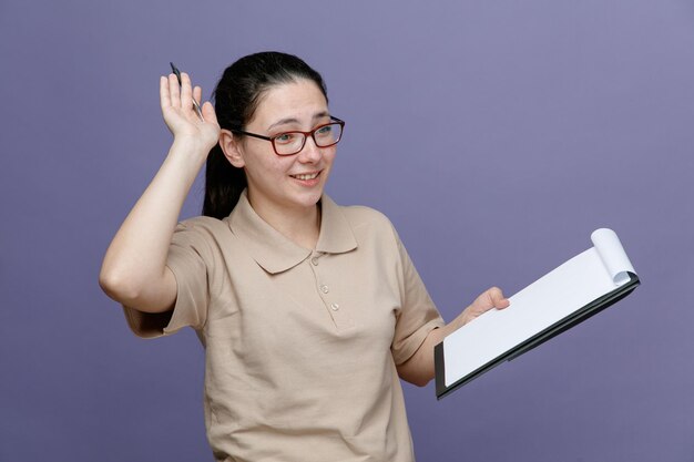Delivery woman employee in blank polo shirt uniform wearing glasses holding clipboard looking aside happy and positive smiling cheerfully standing over blue background