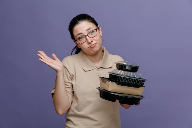 Delivery woman employee in blank polo shirt uniform wearing glasses holding food containers looking confused having doubts raising arm standing over blue background